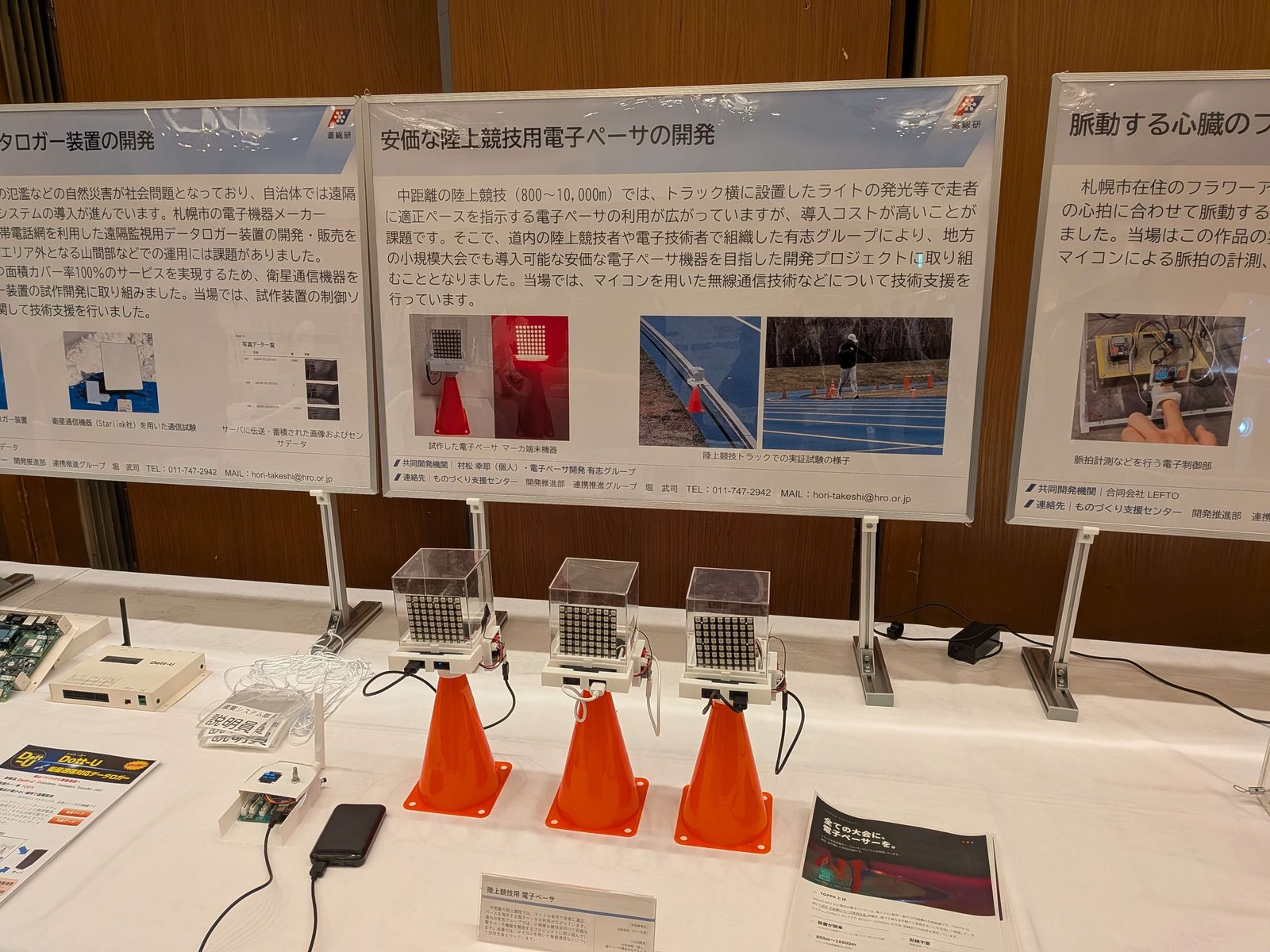 道総研の発表会のブース。白い卓にオレンジのコーンとマーカー3台が並び、奥に「安価な陸上競技用電子ペーサの開発」のポスターが掲示されている