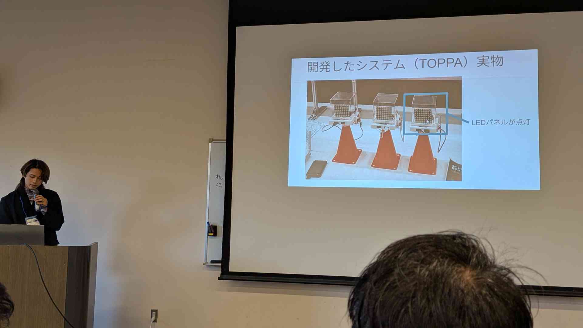 オープンソースカンファレンス北海道の会場。左手前で登壇しており、右のスクリーンに「開発したシステム（TOPPA）実物」のスライドが映っている